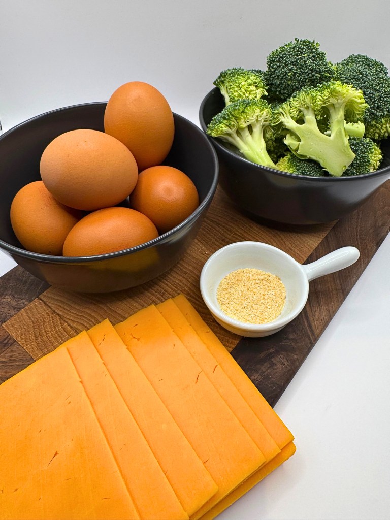 The ingredients to the crustless broccoli cheddar quiche are laid out on a dark brown cutting board. 5 eggs in a bowl, cut broccoli in a bowl, garlic powder in a smaller bowl, and slices of cheddar cheese.