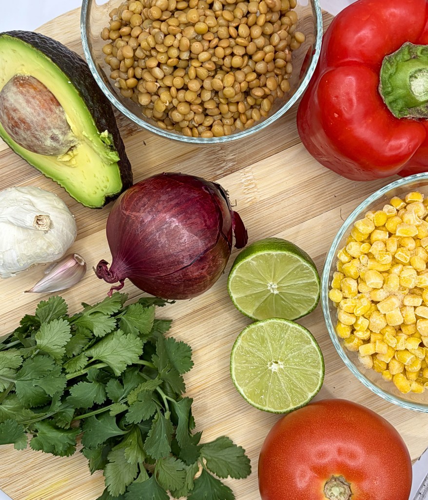 The ingredients for this lentil and corn chopped salad are layed out on a wooden cutting board. Lentils and corn in separate bowls, a lime cut in half, an onion, garlic head, bunch of cilantro, tomato, pepper and avocado laid out.