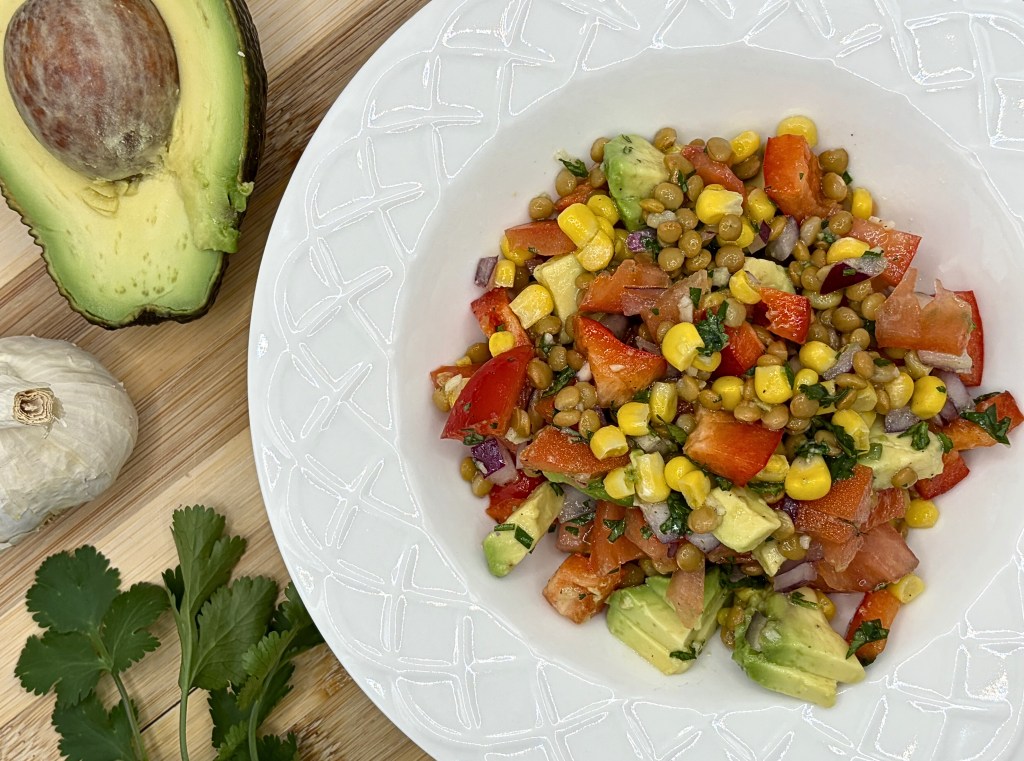 The Lentil & Corn Chopped Salad is in a white bowl in the right center of the photo. Cilantro, garlic and avocado are around the outside of the dish.